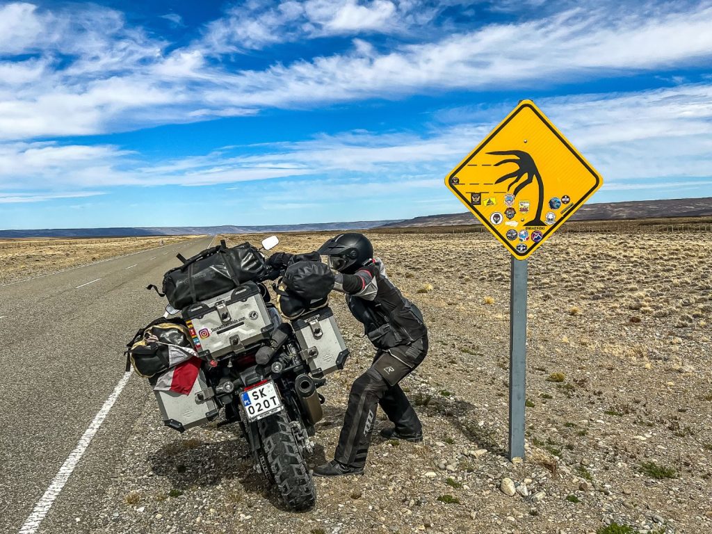 Pretending to hold up a motorcycle in strong winds next to a roadside warning about it.