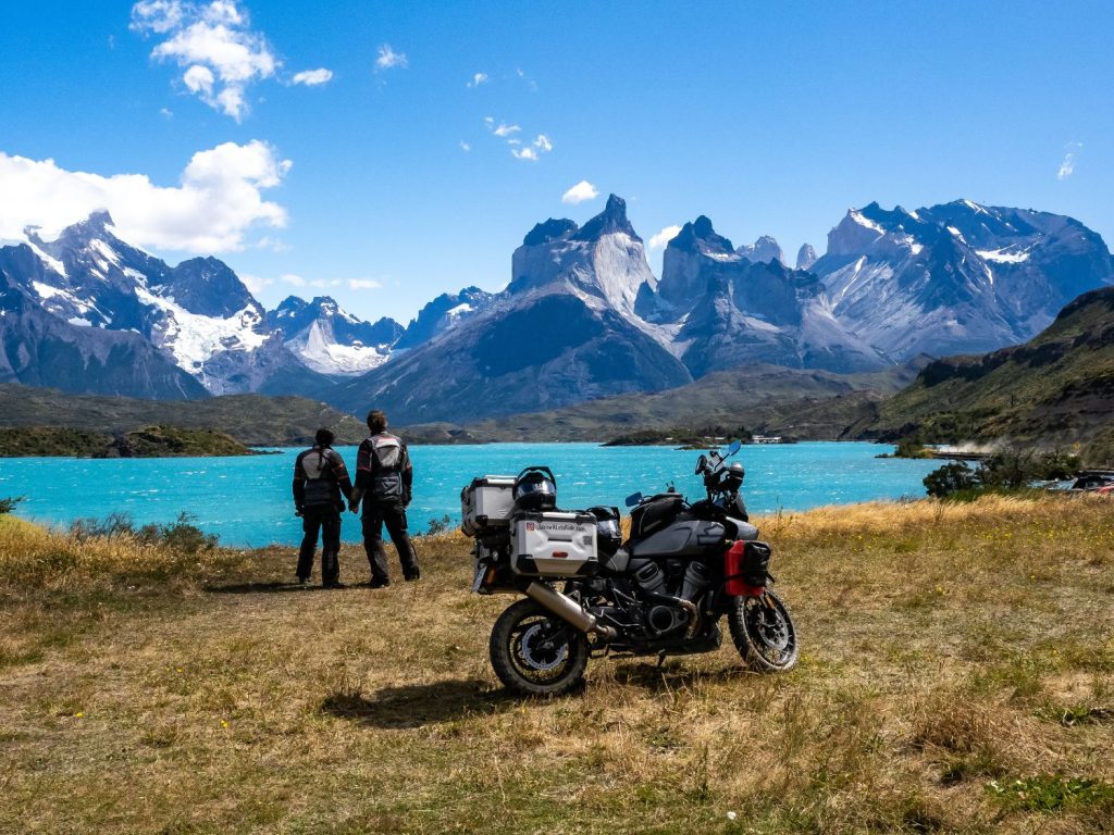 Ela and Greg Kedzior holding hands near a motorcycle admiring the view of mountains and a lake.