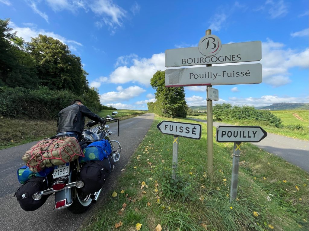 A man sitting on his motorbike, next to the "Pouilly-Fuisse" sign in France.