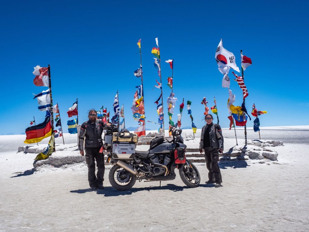 Ela and Greg Kedzior standing with a motorcycle and lots of national flags on the Bolivian salt flats