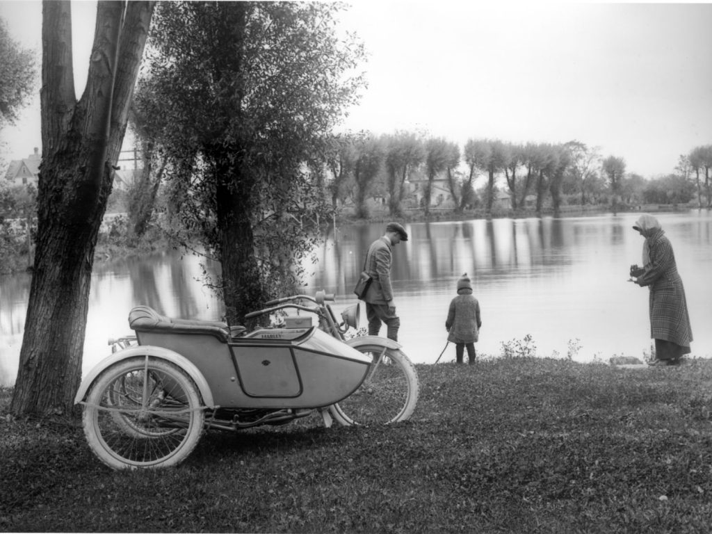The image captured on a lantern slide. It shows a man, woman and child standing by a lake with a Harley-Davidson motorcycle and sidecar in the foreground.